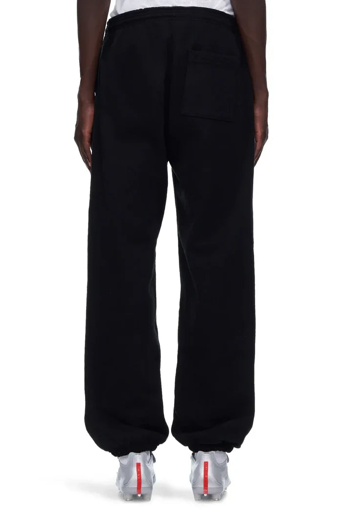 A person faces away wearing 100% Cotton Sp5der Sweatpants—Unisex Black Jogger Pants with "I Love Sp5der" graphic and a relaxed fit—paired with white sneakers featuring red accents against a plain white background.
