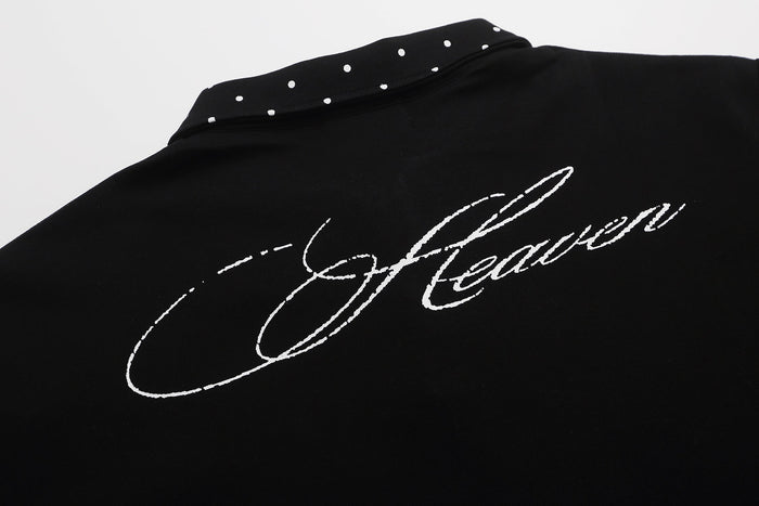 Black graphic polo shirt with white script print and a studded polka dot collar on a clean background
