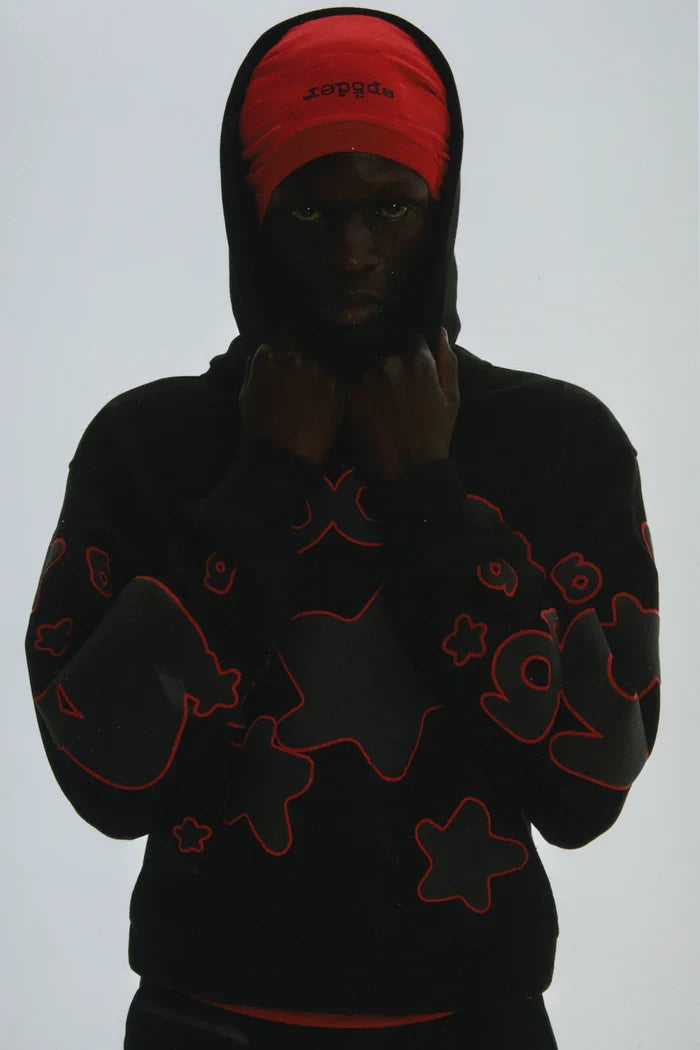 A person in the Sp5der 440GSM Heavyweight Star Hoodie – Black & Grey Oversized Streetwear Pullover stands against a plain background, hood up over a partially hidden red text cap, showcasing bold star designs.