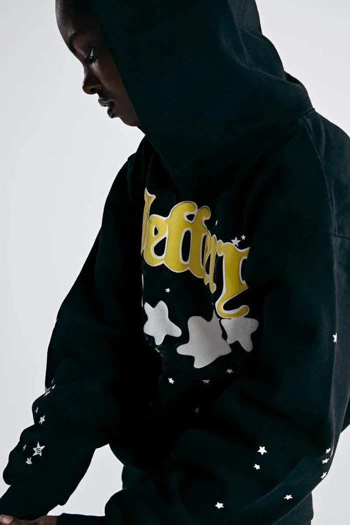 A person wearing the Sp5der Jeffery Hoodie – Black Web 440GSM Heavyweight Streetwear Pullover with yellow lettering and white star and cloud designs looks downward against a plain, light background.