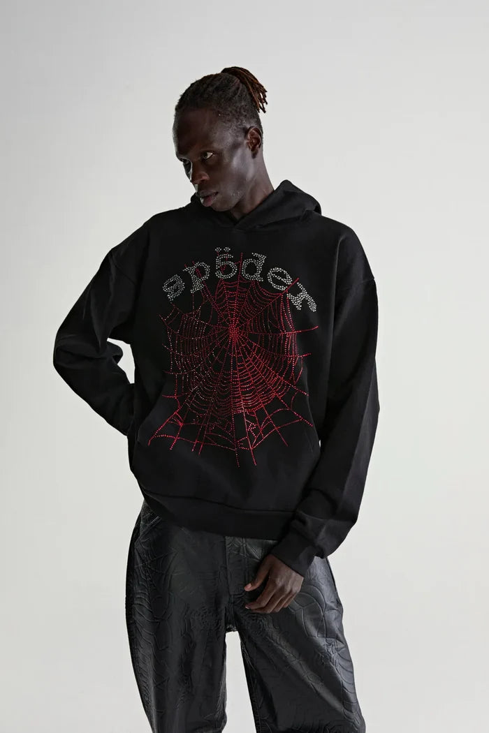 A person in a Sp5der Rhinestone Web Hoodie – Black 440GSM Heavyweight Oversized Streetwear Pullover, with a red spiderweb graphic and stylized "spider" text, stands with one hand on their hip against a light background.