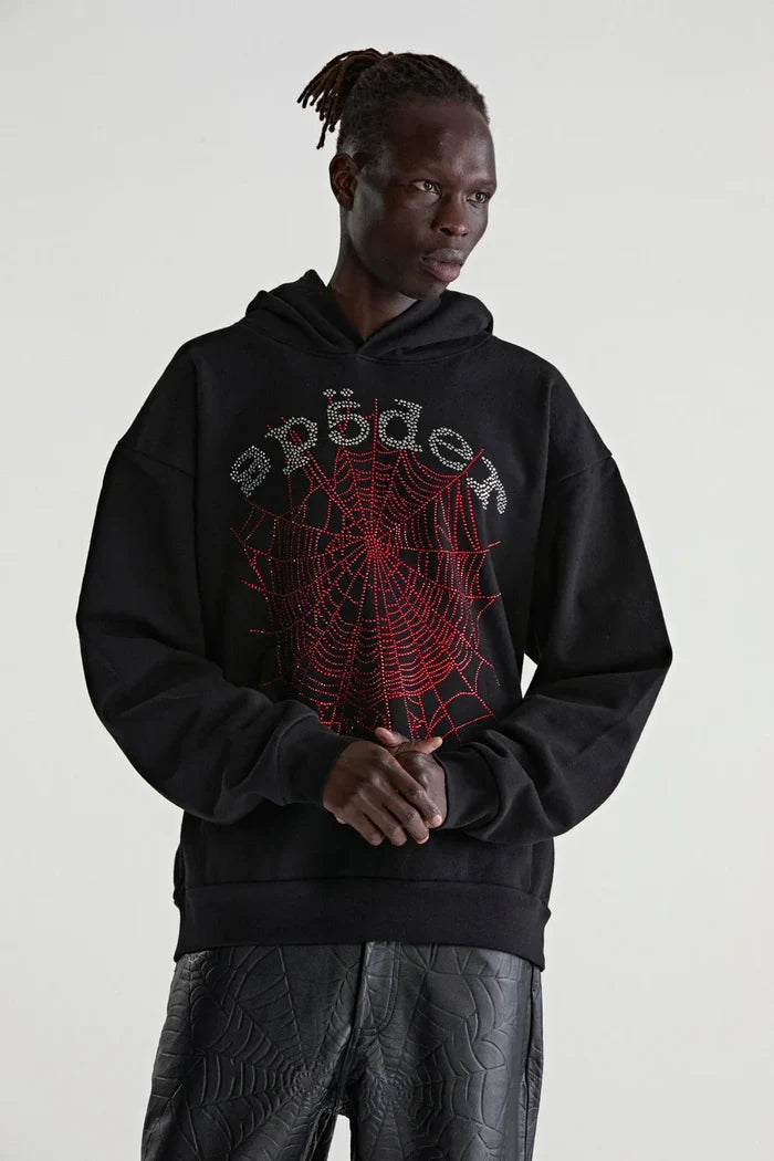 Wearing a Sp5der Rhinestone Web Hoodie – Black 440GSM Heavyweight Oversized Streetwear Pullover with black spiderweb-patterned pants, a person stands before a plain light backdrop, embodying luxury streetwear style.