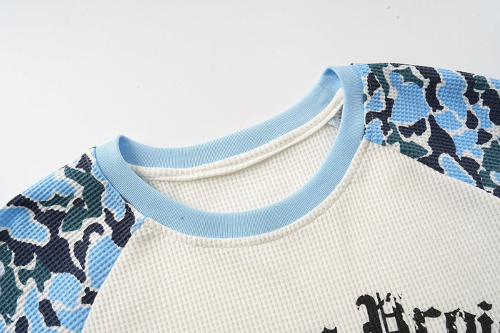 Close-up of a camo raglan graphic tee with light blue ribbed collar and textured cream waffle-knit fabric