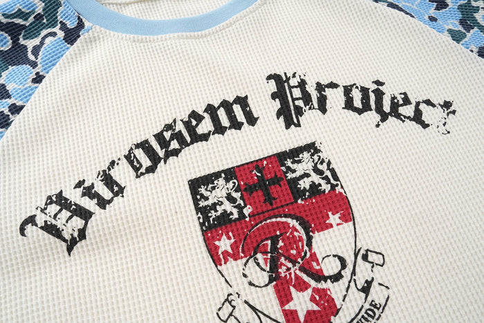 Cream raglan graphic T-shirt with blue camo sleeves and distressed varsity-style print on the chest