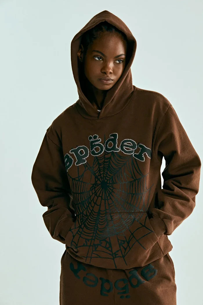 A person in the Sp5der Web Hoodie – Mocha Brown 440GSM Heavyweight Streetwear Pullover and matching pants with spɪder text and a large spiderweb graphic on the front embodies bold streetwear style, standing hands in pockets.