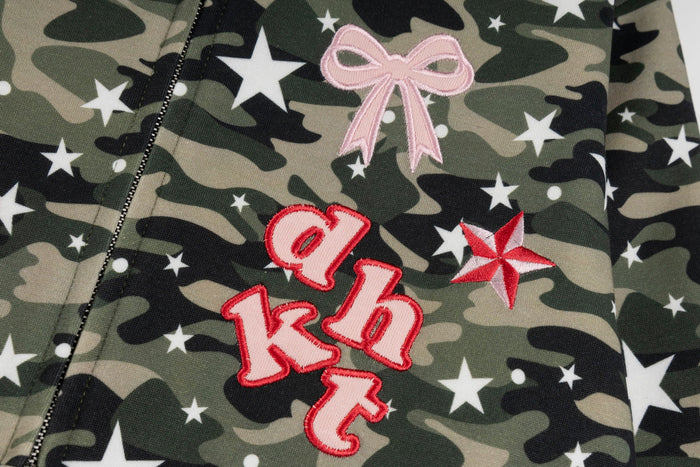 Close-up of front chest embroidery on camo zip hoodie with pink bow, lettering, and star patch