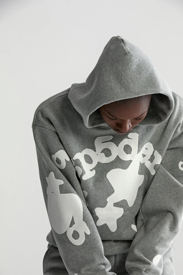 A person in the Sp5der Stars Hoodie – Grey 440GSM Heavyweight Streetwear Pullover, featuring bold white graphics and text, looks downward with the hood up against a plain light background, highlighting its comfort and style.