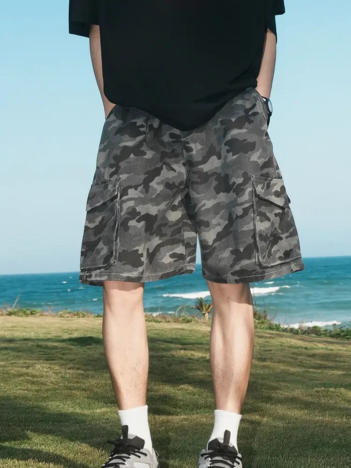 A person in Men's Dark Grey Camo Cargo Shorts with Elastic Waist and Flap Pockets, a black shirt, white socks, and sneakers stands on grass by the sea under a blue sky.