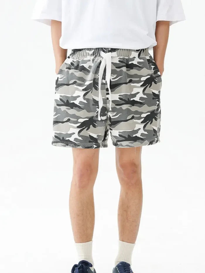 A person stands with hands in pockets against a plain white background, wearing a white t-shirt, navy sneakers, and Men's Urban Camo Cotton Drawstring Shorts - Gray Camouflage Relaxed Fit with a white drawstring.