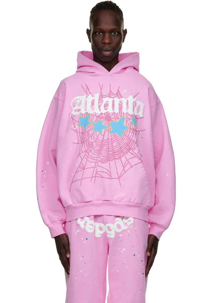A person models the Sp5der Atlanta Hoodie – Pink 440GSM Heavyweight Y2K Streetwear Pullover, featuring a white web design, blue and yellow stars, and text, paired with matching pants against a plain background.