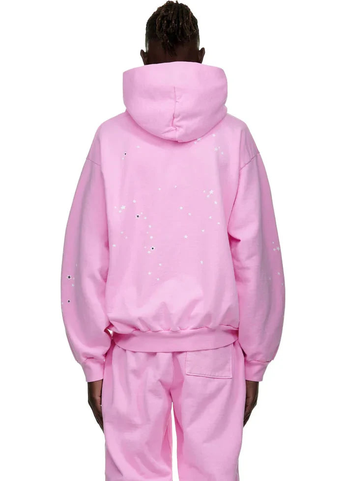 A person faces away from the camera in a Sp5der Atlanta Hoodie – Pink 440GSM Heavyweight Y2K Streetwear Pullover with matching pants. The hood is up, and the hoodie features white paint splatter details on the back and sleeves.