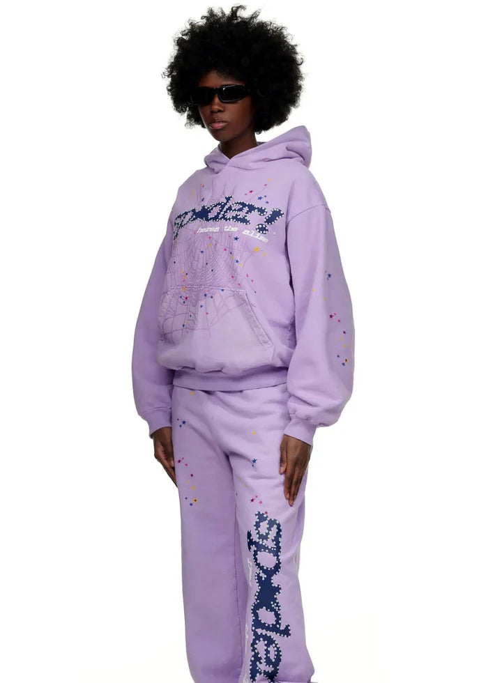 A person sports the Sp5der “Hares the Slime” lavender purple hoodie—440GSM heavyweight fleece with spider-web text and graphics—styled with matching sweatpants, an afro, black sunglasses, and vibrant streetwear vibes.