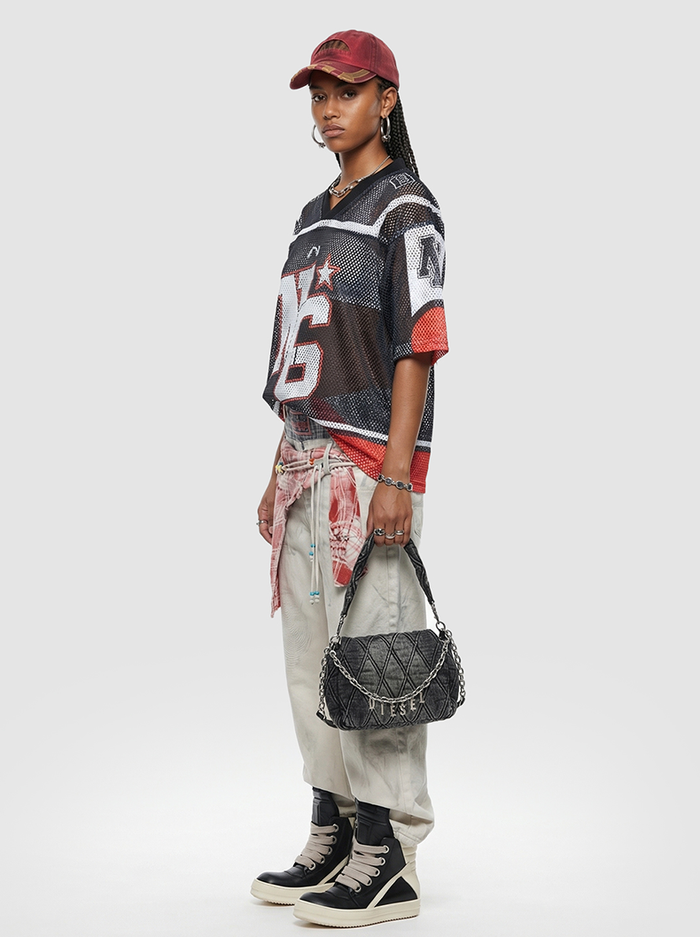 Model wearing a black mesh jersey T-shirt with bold graphic print, styled with cream pants, red cap, and black sneakers