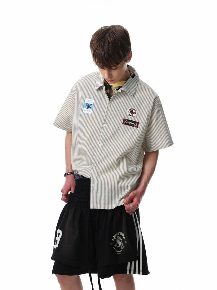 Beige pinstripe short-sleeve button-up with graphic patches, styled on a model with black streetwear shorts