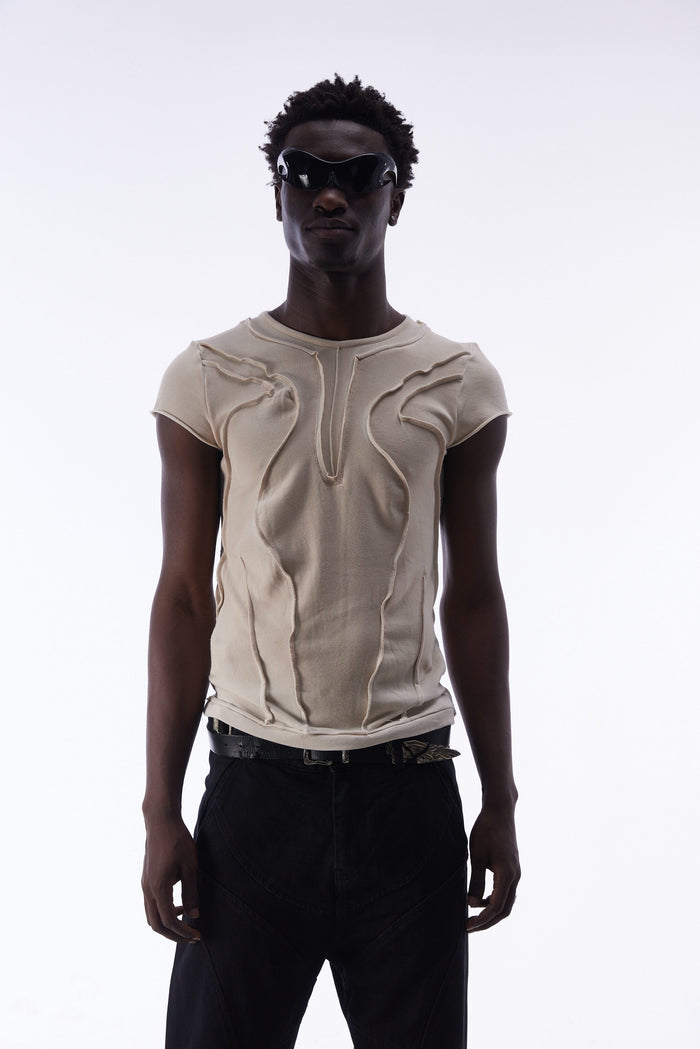 Model wearing a beige sculpted seam cotton tee with curved panel detailing, black trousers, and futuristic sunglasses