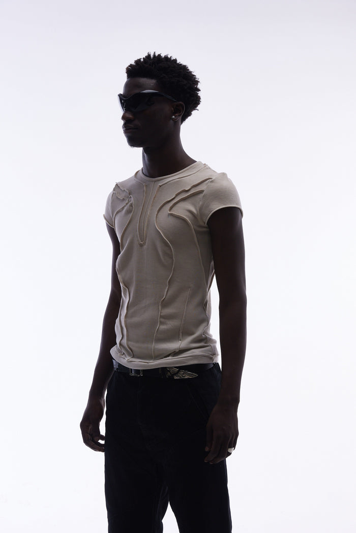 Beige sculpted cotton tee with raw seam paneling, worn by a model in black sunglasses and dark pants