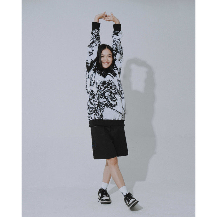 A person poses with arms raised, wearing Seakoff's Premium Skeleton Warrior Knit Sweater—a gothic jacquard design—paired with black shorts, sneakers, and white socks. Their shadow is cast on the light gray wall, enhancing the ensemble's gothic streetwear vibe.