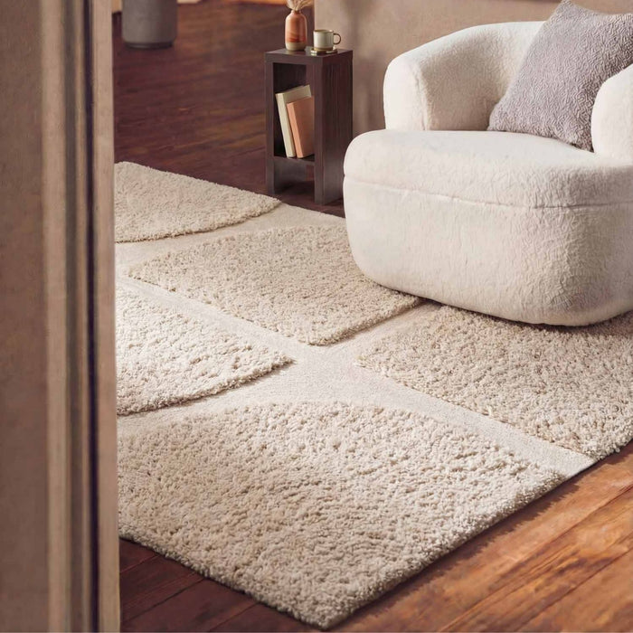 Beige textured rug on a wooden floor with a white armchair in the corner.