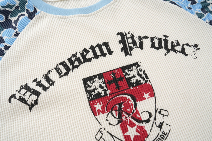 Close-up of front crest graphic on cream waffle-knit tee with black distressed text and red shield emblem